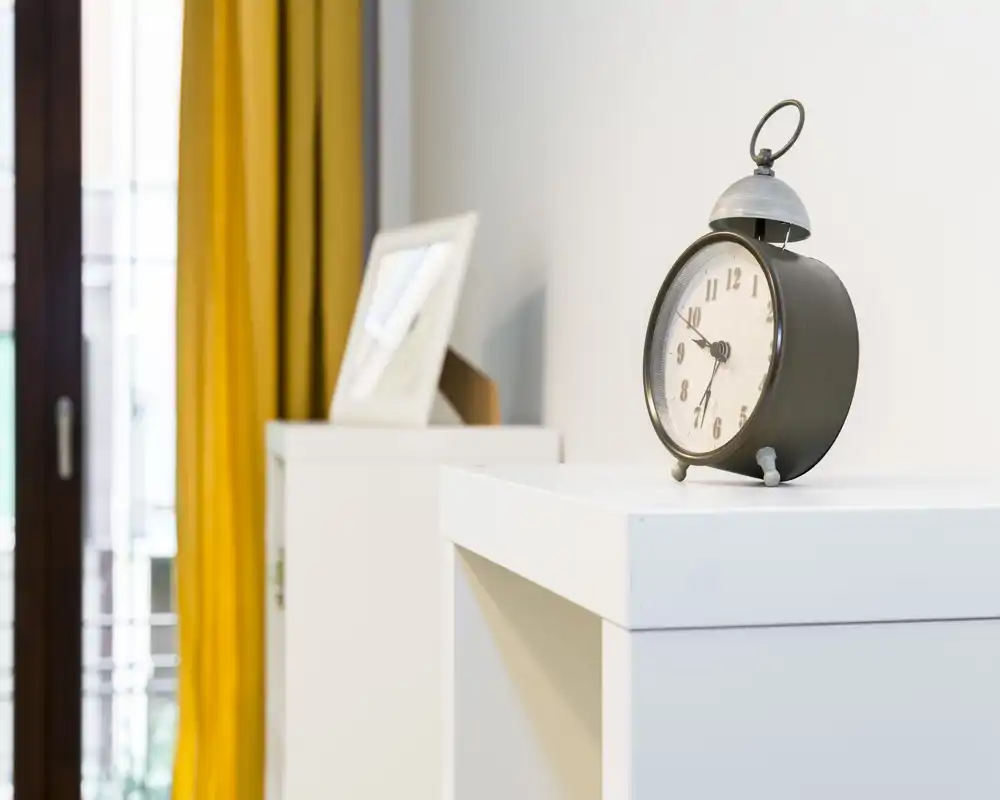 Dettaglio di una camera ben illuminata con un comodino bianco minimalista, una sveglia vintage e luce naturale morbida proveniente da una finestra con tende gialle.