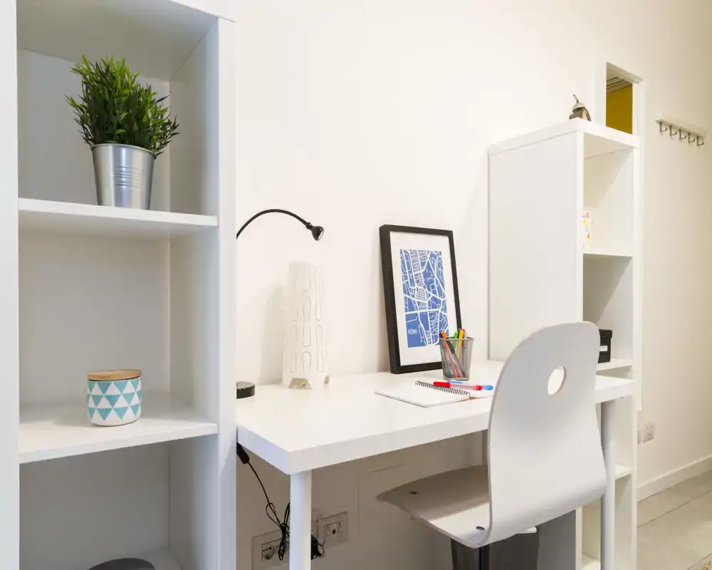 Angolo studio luminoso e minimale con scrivania bianca, sedia, stampa incorniciata e scaffalatura — ideale per un piccolo home office.