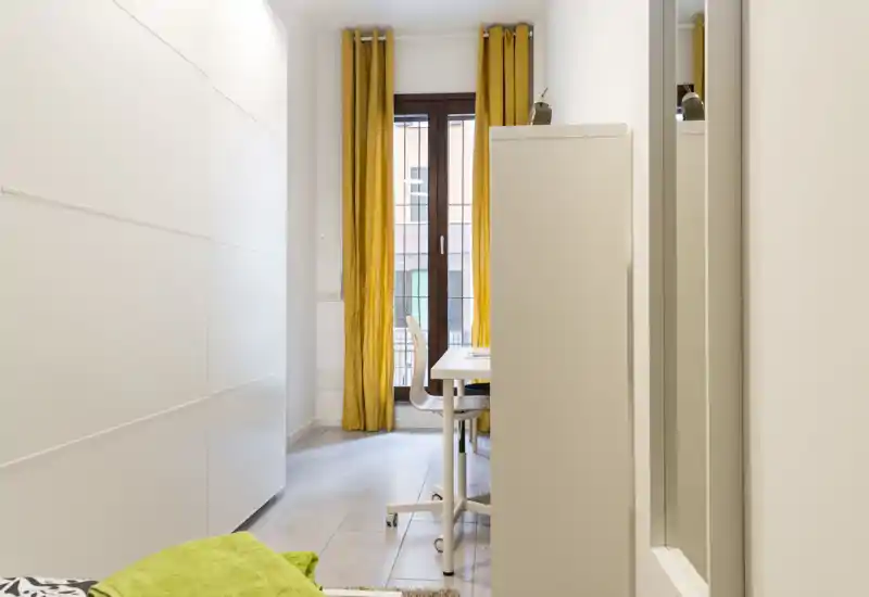 Angolo camera compatto con parte del letto visibile e una postazione vicino alla finestra con tende gialle; ambiente pulito, luminoso e funzionale, adatto a una persona.