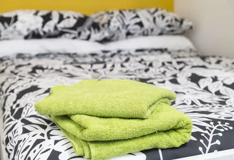 Camera luminosa su un letto ben fatto con copripiumino a motivo e asciugamani verdi piegati sopra, per un aspetto fresco e accogliente.