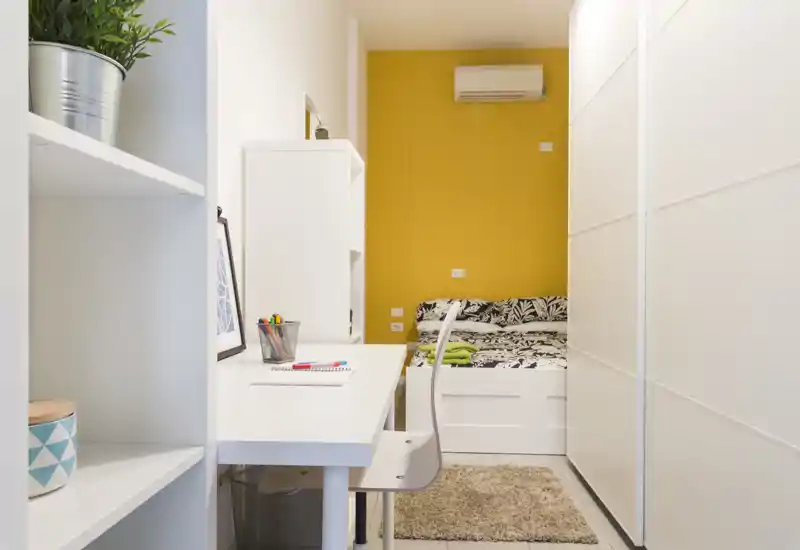 Camera da letto compatta e ben arredata con una scrivania bianca in primo piano e un letto ben rifatto davanti a una parete d'accento gialla. L'ambiente ha mobili moderni, condizionatore sopra il letto e tessili morbidi che rendono la stanza accogliente.