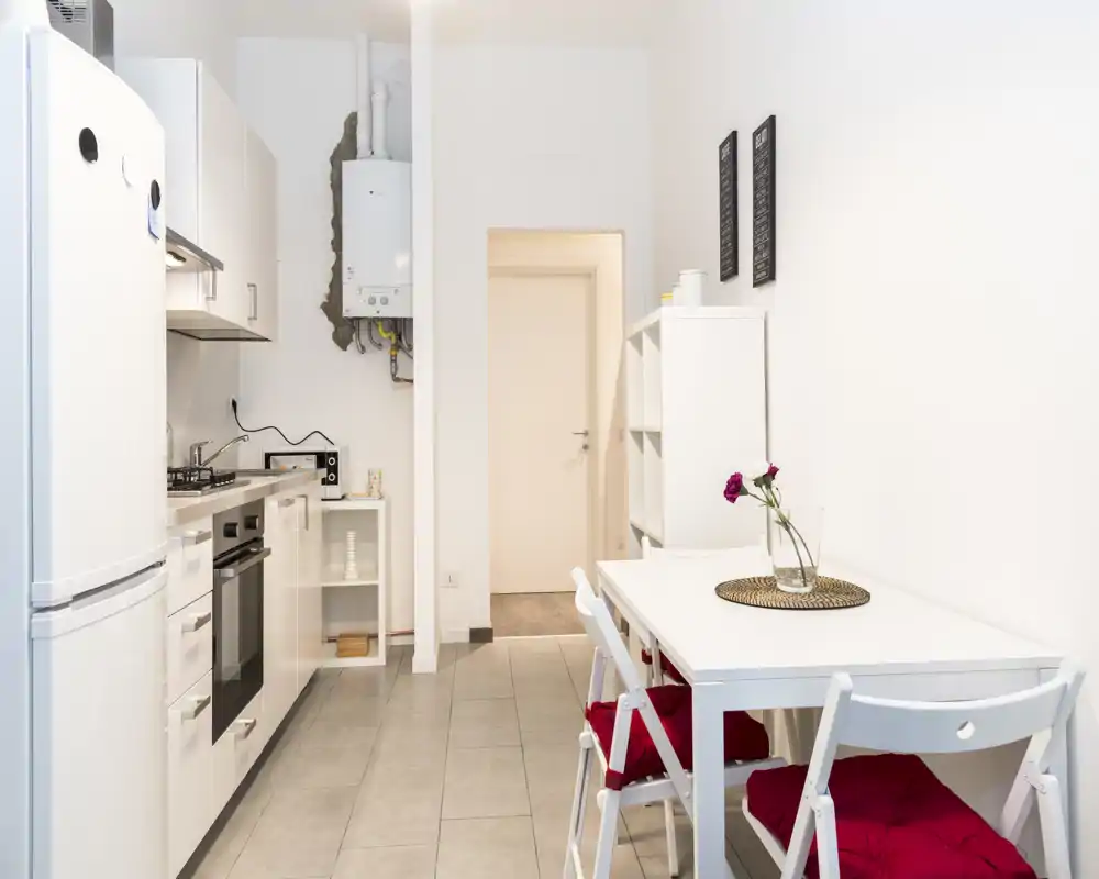 Cucina compatta e luminosa con elettrodomestici integrati e piccolo tavolo da pranzo. Pensili bianchi e arredo minimale per un ambiente pulito e arioso, perfetto per un appartamento moderno.