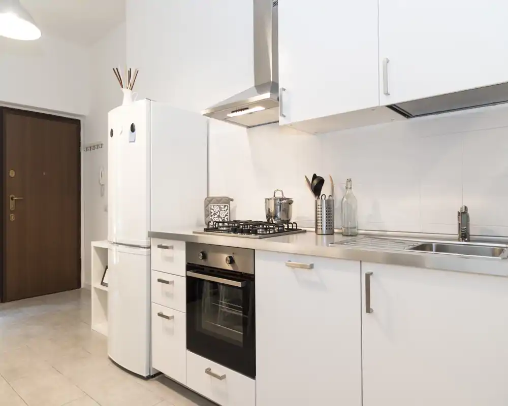 Cucina moderna e compatta con mobili bianchi, top in acciaio inox, forno e piano cottura a gas integrati; ambiente luminoso e pulito, ideale per affitti brevi.
