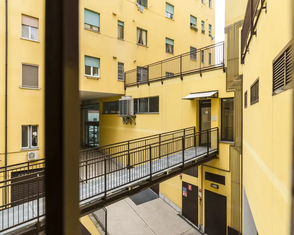 Cortile interno luminoso con passerella pedonale sopraelevata, facciate gialle e ringhiere metalliche, perfetto per descrivere le aree comuni di un appartamento in città.