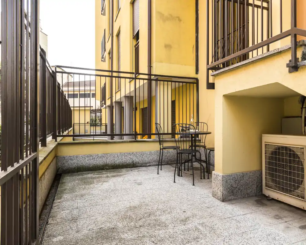 Terrazza privata con ringhiere in metallo e un piccolo tavolo con sedie, ideale per rilassarsi o cenare all'aperto. Pavimentazione in pietra e unità esterna del condizionatore, con vista sugli edifici gialli adiacenti.