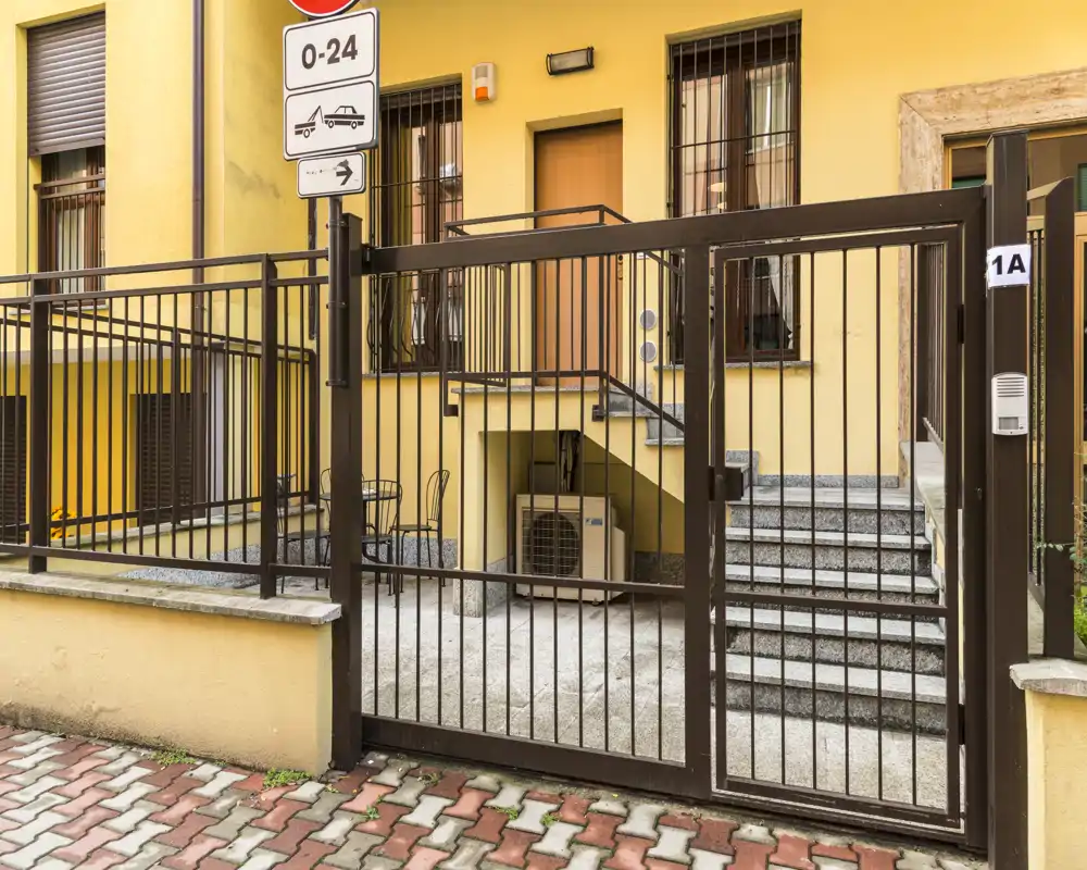 Cortile d'ingresso recintato con gradini che conducono alla porta dell'edificio; cancello metallico, citofono e segnaletica ben visibili.