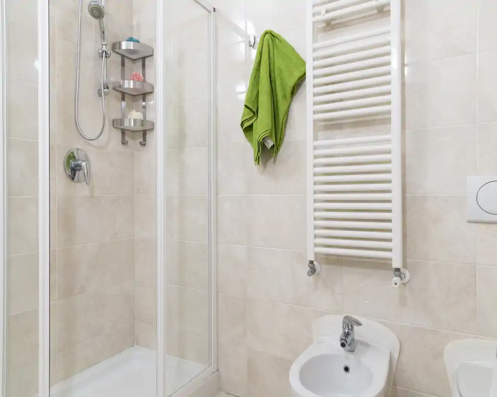 Bagno moderno e luminoso con box doccia in vetro angolare, bidet, termoarredo e piastrelle neutre — ben illuminato e pulito.