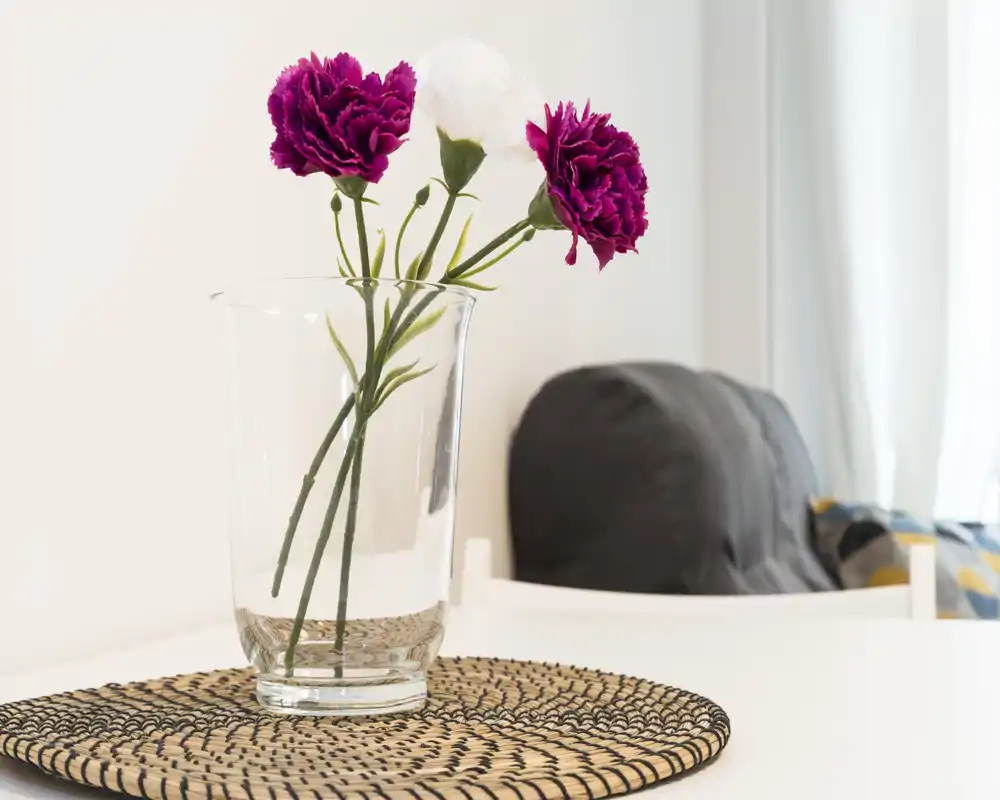 Dettaglio di un soggiorno luminoso con un vaso di vetro trasparente e fiori su un tavolo bianco, luce naturale soffusa e una poltrona sfocata sullo sfondo.