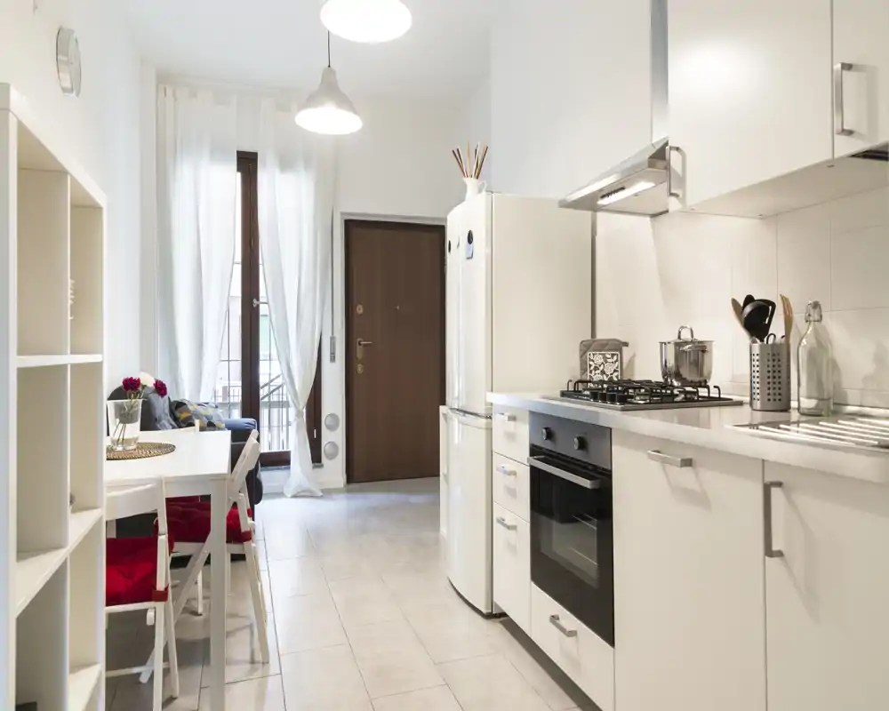 Cucina moderna e luminosa con forno integrato, piano cottura a gas e un piccolo angolo pranzo accanto alla finestra-porta che lascia entrare luce naturale.