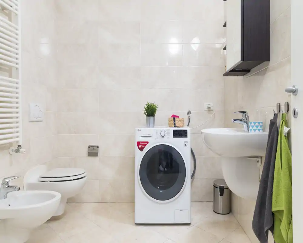 Bagno moderno e luminoso con WC sospeso, bidet, lavabo e una lavatrice posizionata tra i sanitari; piastrelle neutre e accessori ordinati.