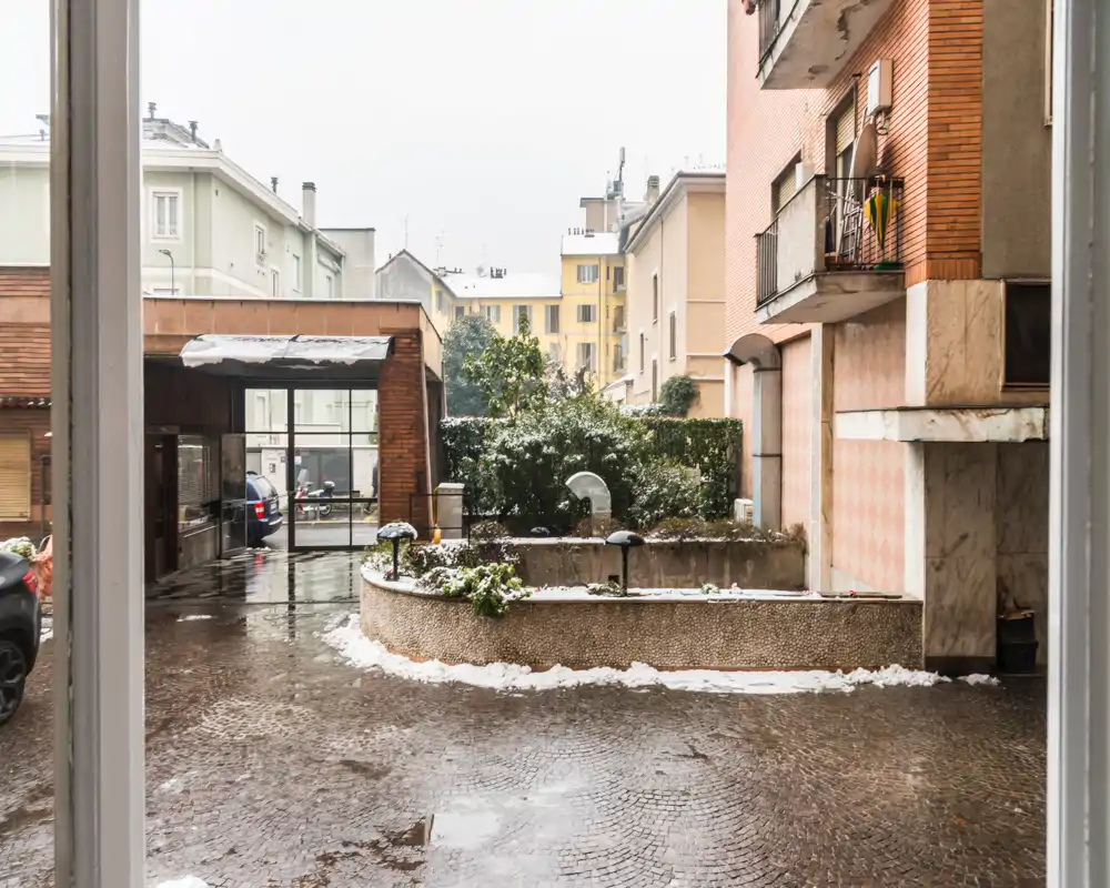 View of a residential courtyard and entrance area with wet cobbled pavement, planters, parked cars and surrounding apartment façades — useful exterior shot showing access and immediate outdoor space.