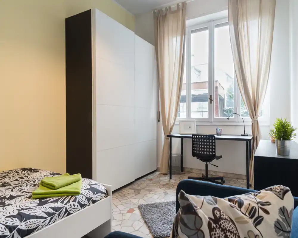 Bright, neatly styled bedroom with a single bed, wardrobe and a desk by the window—ideal for a student or professional. Natural light and neutral tones create a calm, welcoming atmosphere.