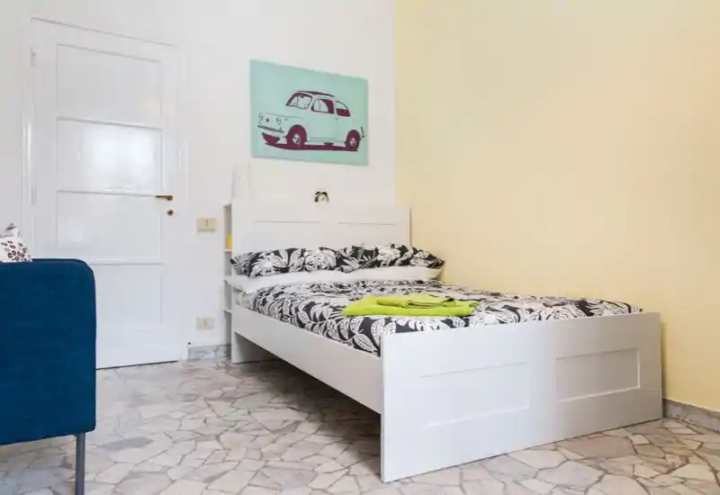 Bright, neatly styled bedroom with a white bed frame, patterned bedding and a cheerful wall print. The room features tiled flooring and clean, minimal decor ideal for short-term rentals.