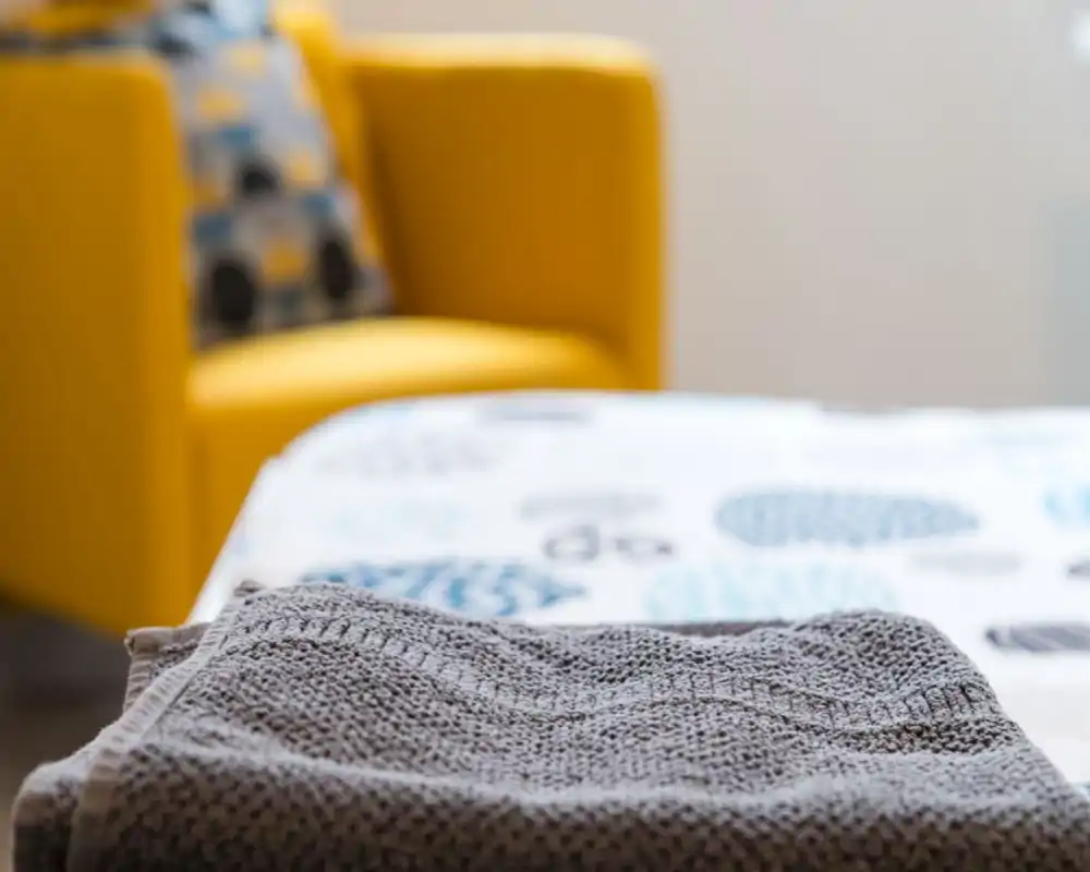Plano detalle de ropa de cama doblada en primer plano y un sillón amarillo brillante al fondo; la imagen destaca la textura de la cama y un rincón acogedor.