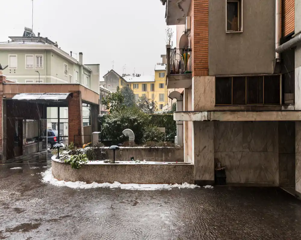 Patio exterior con pavimento de empedrado mojado, jardineras bajas y fachadas de apartamentos alrededor; tiempo nublado con ligera nieve en los bordes.