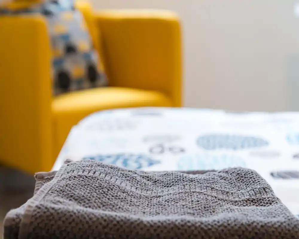 Primer plano de una toalla doblada sobre la cama con un cubrecama estampado y un sillón amarillo de fondo, destacando los textiles y una puesta en escena acogedora.