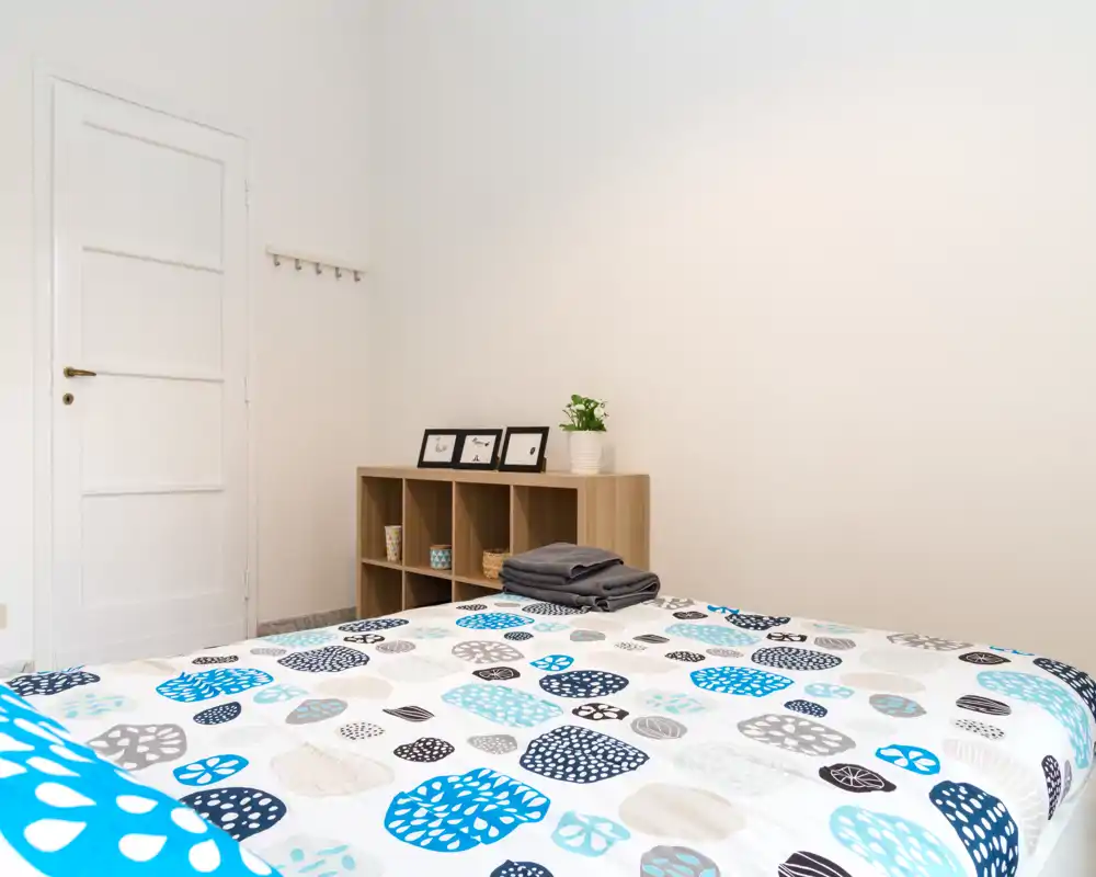 Dormitorio luminoso y minimalista con edredón estampado, estantería abierta y ropa doblada — espacio limpio y bien iluminado, ideal para anuncios.