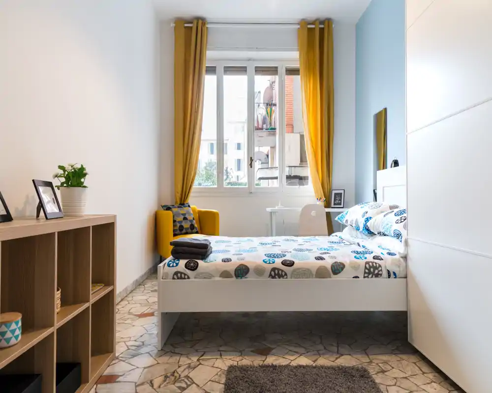 Dormitorio luminoso y bien decorado con cama doble, ropa de cama estampada, un sillón amarillo acogedor y una gran ventana que aporta mucha luz natural.