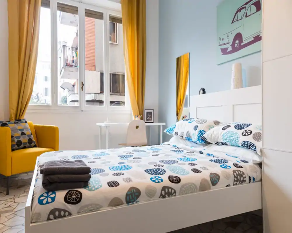 Dormitorio moderno y luminoso con cama doble bien hecha, ropa de cama con estampado, sillón amarillo y grandes ventanas que aportan mucha luz natural.