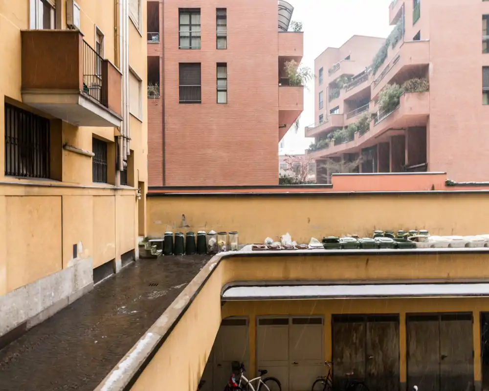 Vista del patio/terraza exterior con fachadas de edificios, macetas y una zona pavimentada; superficies mojadas por la lluvia y bicicletas guardadas abajo.