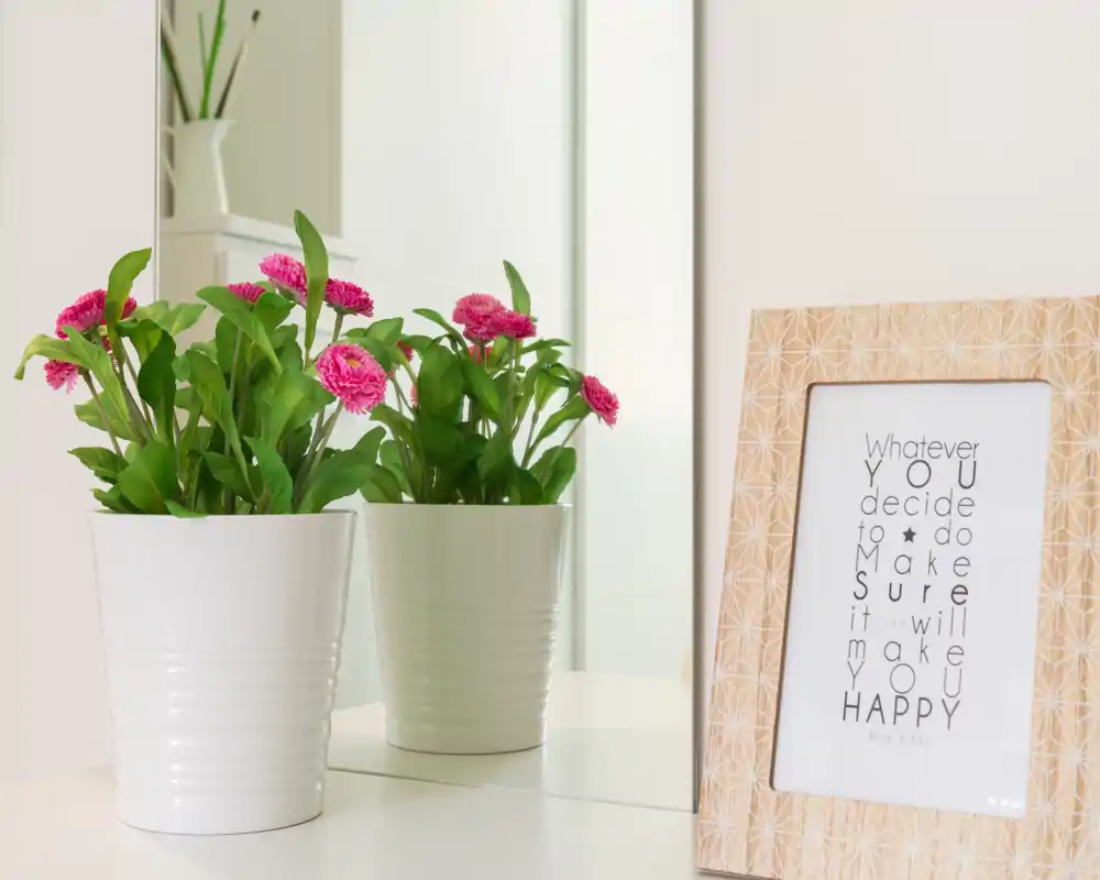 Plano detalle decorativo con una maceta blanca con flores reflejada en un espejo y un marco con una frase en una superficie — imagen pensada para mostrar detalles de decoración del alojamiento.
