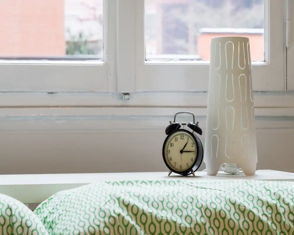 Dormitorio luminoso y minimalista con edredón verde estampado, reloj despertador y lámpara moderna frente a una gran ventana.