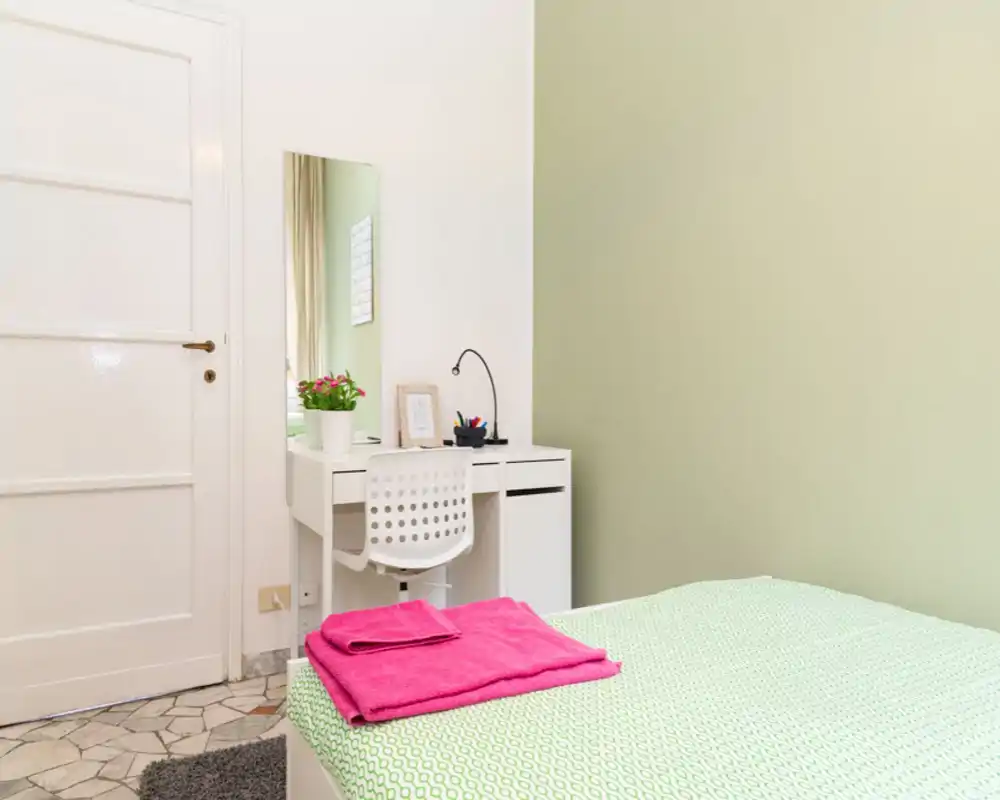 Dormitorio luminoso y ordenado con colcha verde estampada, toallas rosas dobladas y un pequeño escritorio blanco con espejo y planta — ideal para una estancia acogedora.