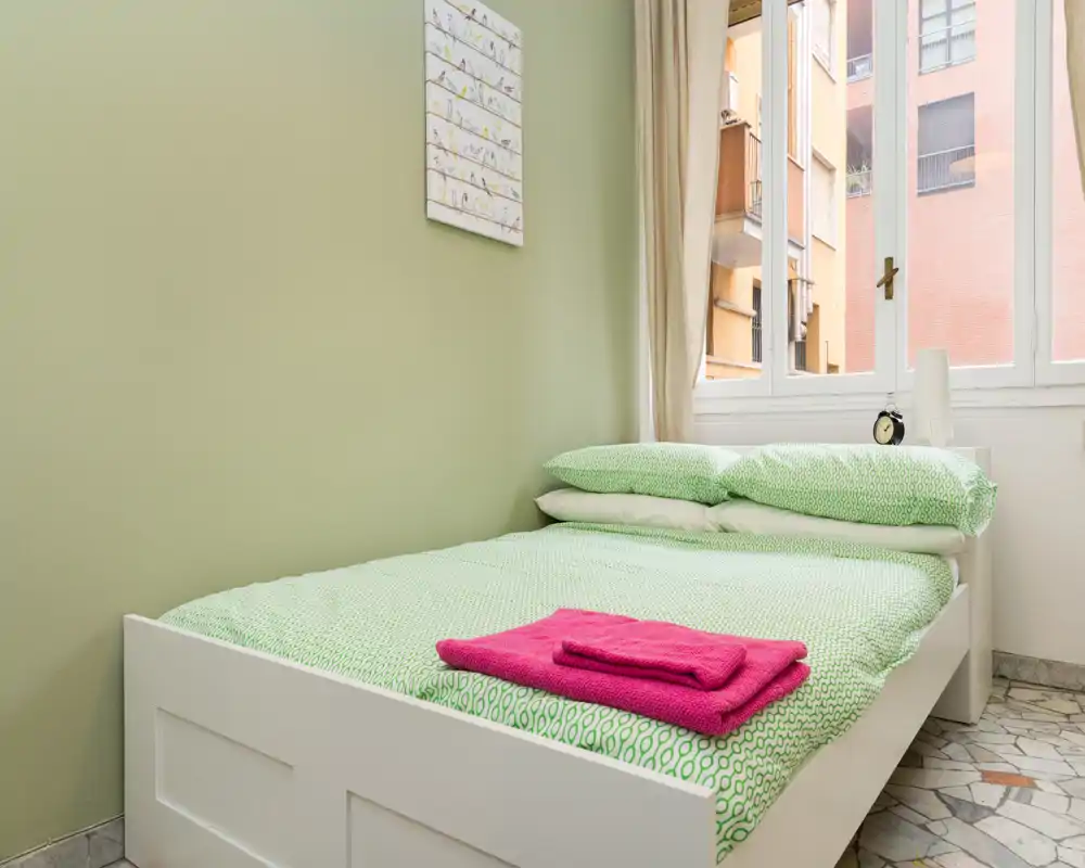 Dormitorio individual luminoso con cama de marco blanco y ropa de cama verde con estampado, toallas rosas dobladas y una gran ventana que aporta luz natural.
