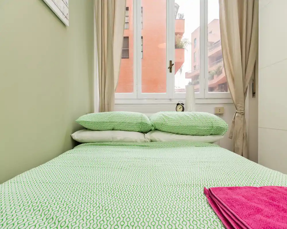 Dormitorio luminoso y ordenado con ropa de cama verde estampada y dos almohadas frente a una gran ventana que aporta luz natural.