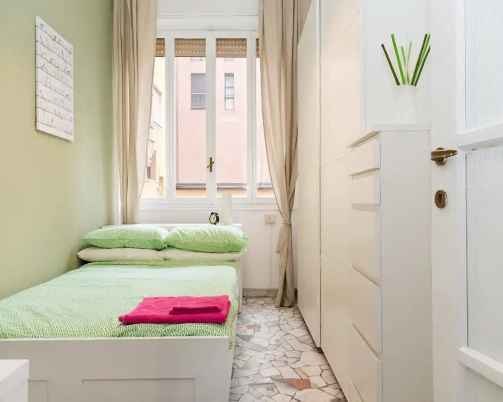Dormitorio luminoso y compacto con una cama individual bien hecha con ropa de cama verde, un armario blanco alto y luz natural desde la ventana.