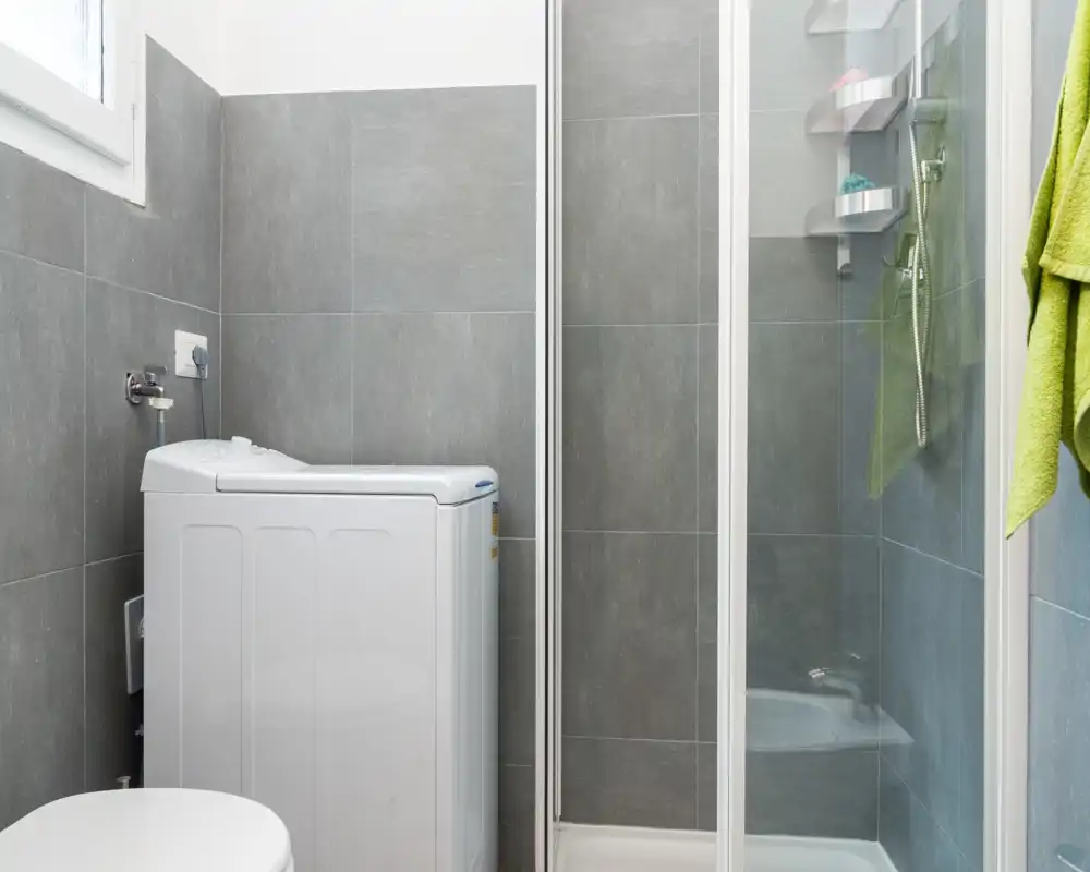 Compact modern bathroom with gray tiled walls, a shower enclosure and a top-loading washing machine; bright, clean and efficiently arranged.