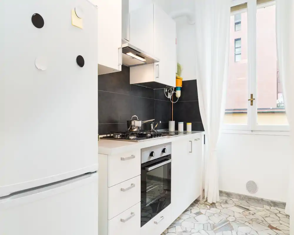 Compact modern kitchen with white cabinetry, built-in oven and gas hob, a fridge at left and a bright window that floods the space with natural light.