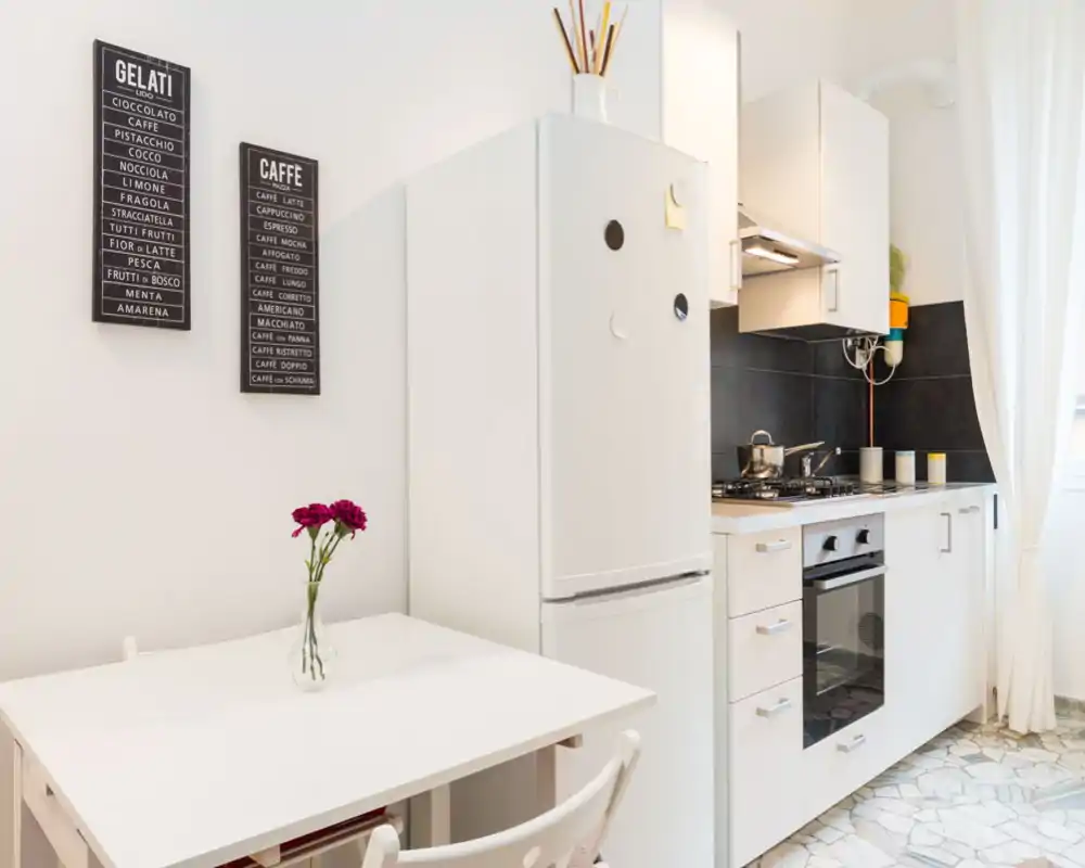 Bright modern kitchenette with white cabinets, gas stove and a compact dining table with a small vase of flowers — tidy and well-lit space ideal for short-term stays.