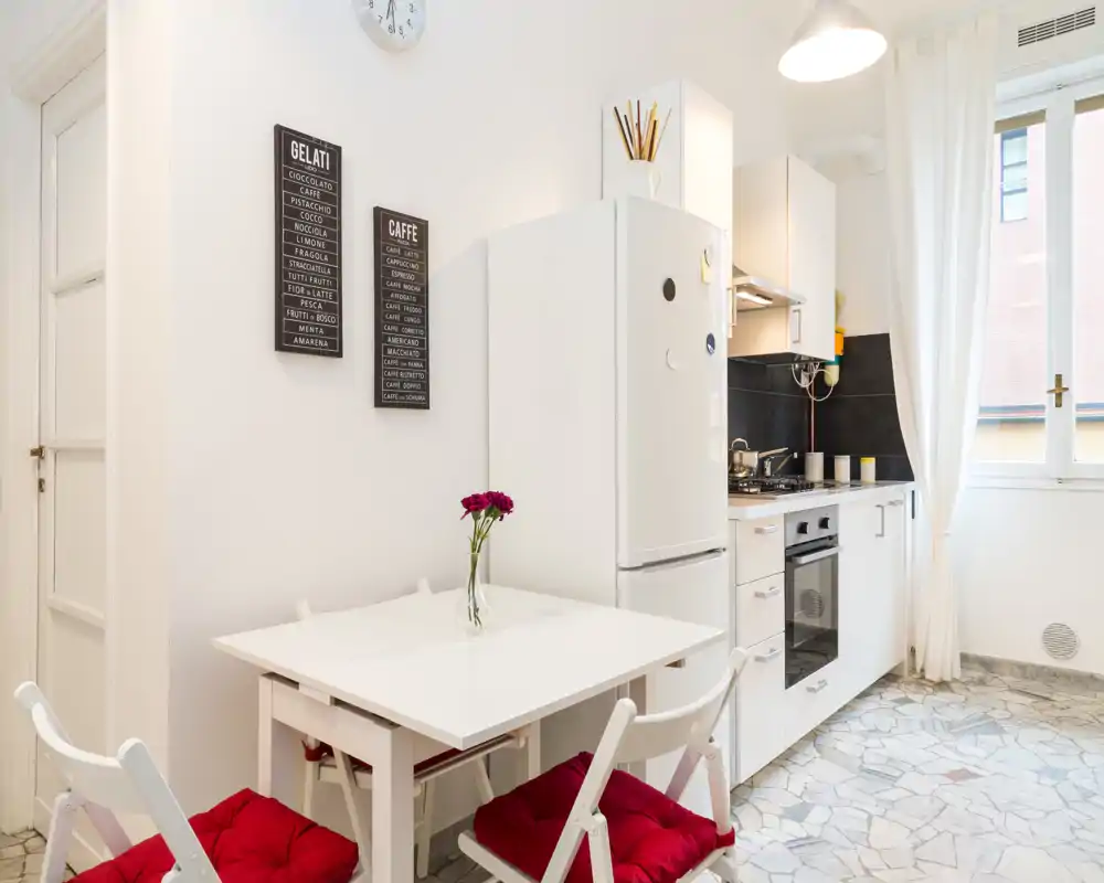 Bright modern kitchen with a compact dining table, white cabinets, refrigerator and oven; a vase with fresh flowers and red seat cushions add a cozy touch.