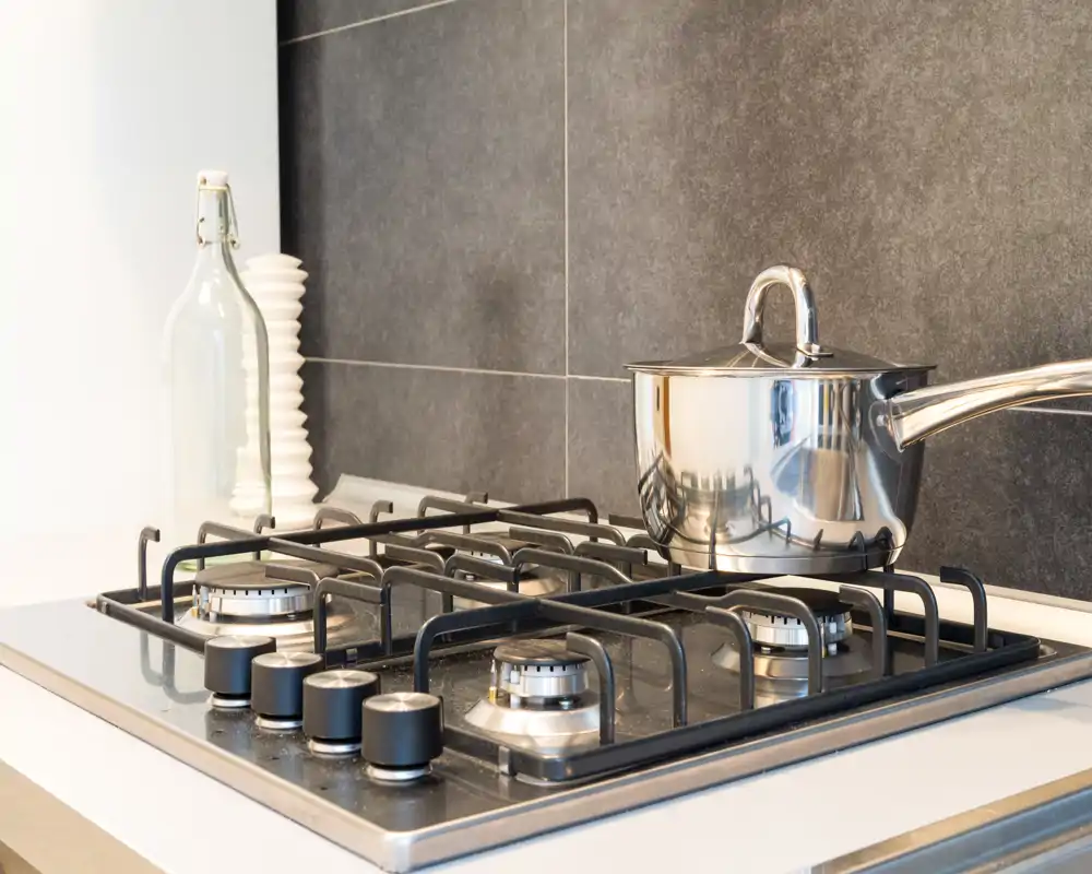 Modern kitchen stovetop with a stainless-steel saucepan, visible burners and control knobs, set against a dark tiled backsplash — bright, clean and well-styled for listing photos.