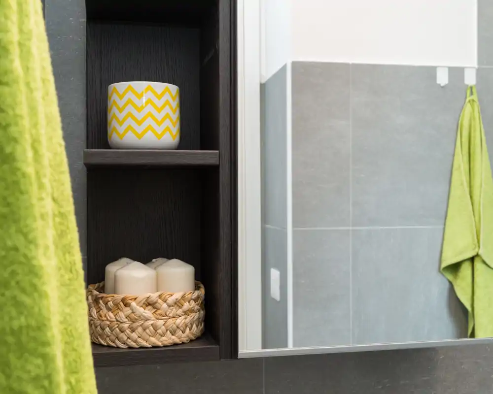 Close-up of a bathroom shelving niche with decorative candles, a patterned cup and green towels visible beside grey tiled walls.