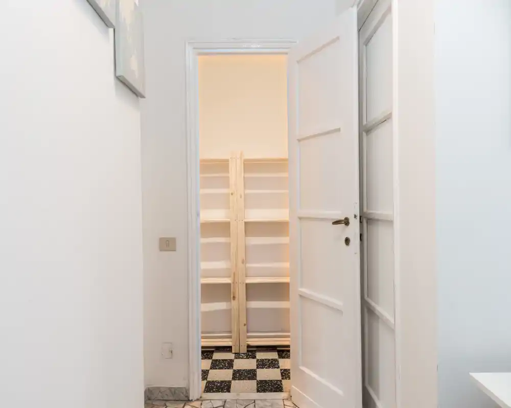 Simple entrance view with an open white door leading to a small shelved storage area; clean walls and tiled floor create a neutral, functional arrival space.