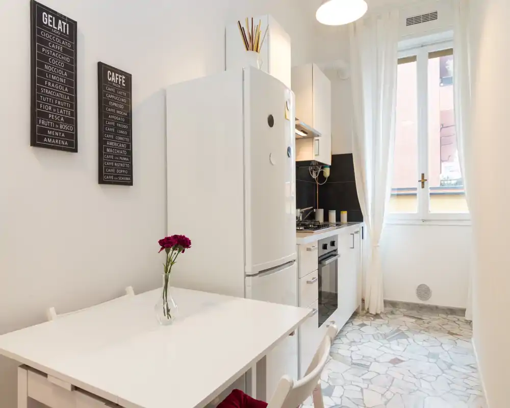 Bright, compact kitchen with white cabinets, a freestanding refrigerator, built-in oven and a small dining table; tasteful decor and natural light from the window create a welcoming space.