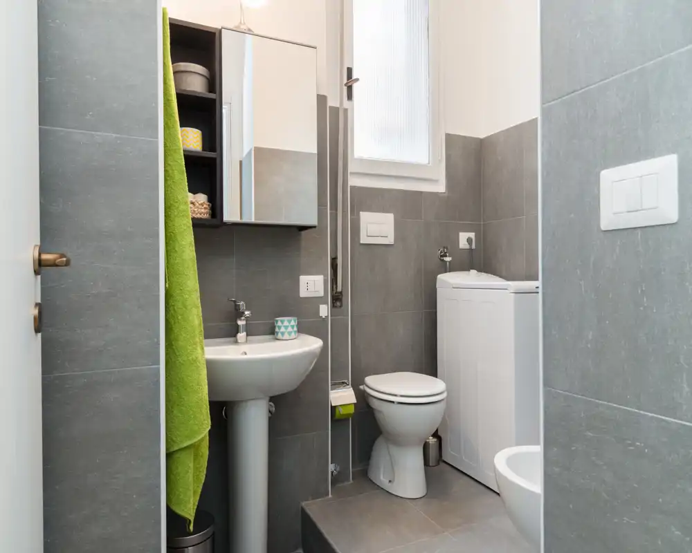 Compact modern bathroom with grey tiles, pedestal sink, toilet and a washing machine. Bright, tidy space with a wall cabinet and mirror above the sink.