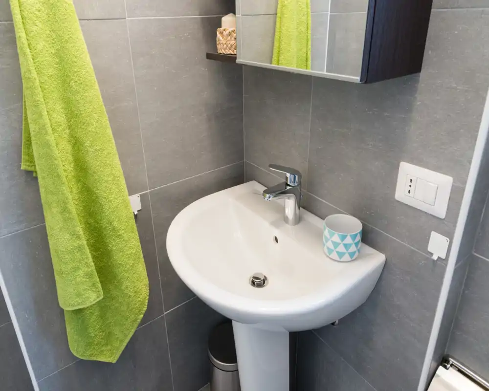 Compact modern bathroom corner with a white pedestal sink, chrome tap, grey tiled walls and a green towel—bright and well-maintained.