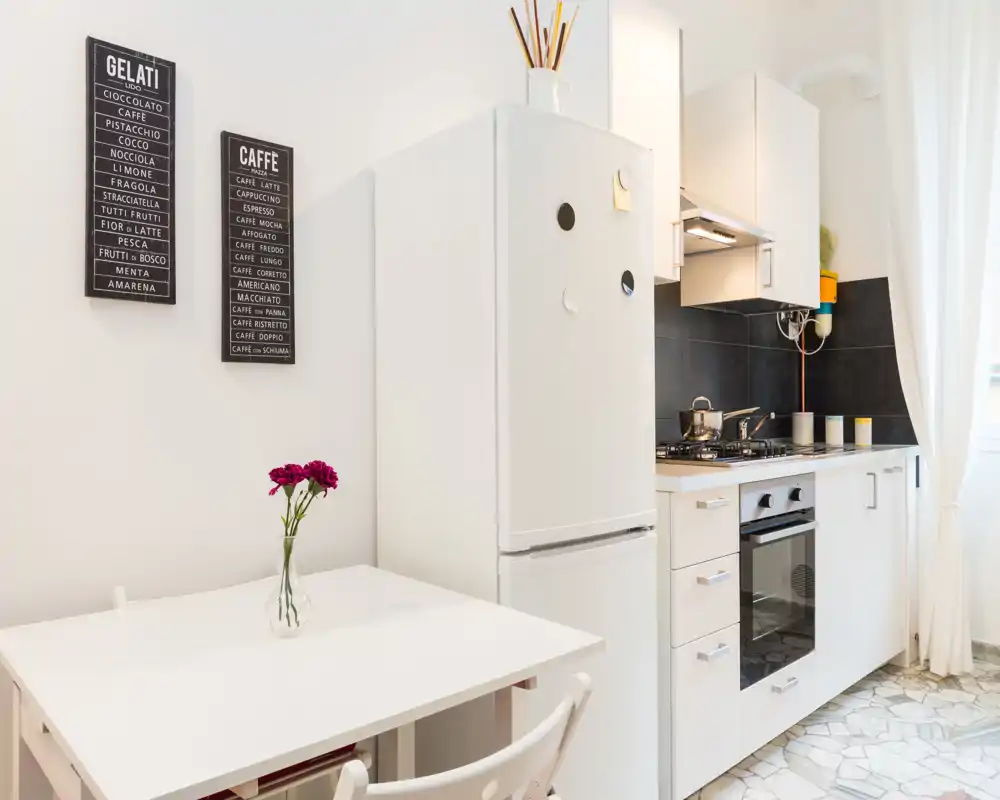 Compact, modern kitchen with a white fridge, built-in oven and gas hob, neat cabinetry and a small dining table with fresh flowers.