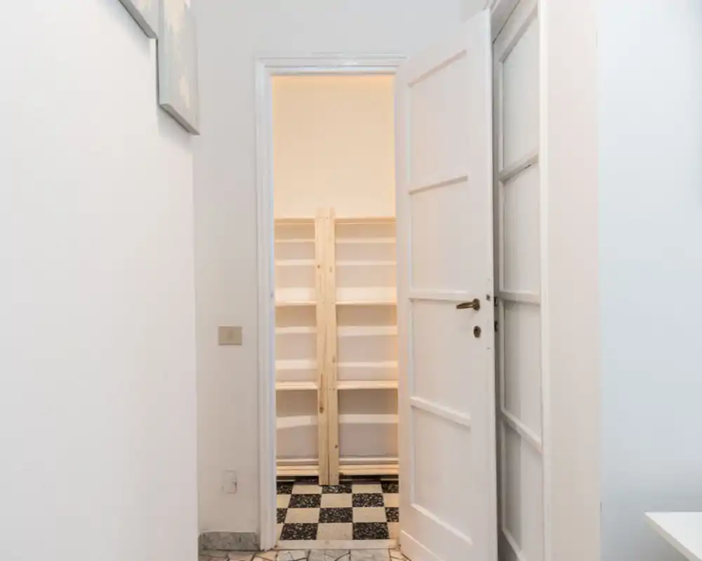 Bright, minimal hallway/entrance view with an open door leading to a small storage area or closet with shelving. Neutral white walls and checkerboard floor tiles create a clean, simple entrance space.