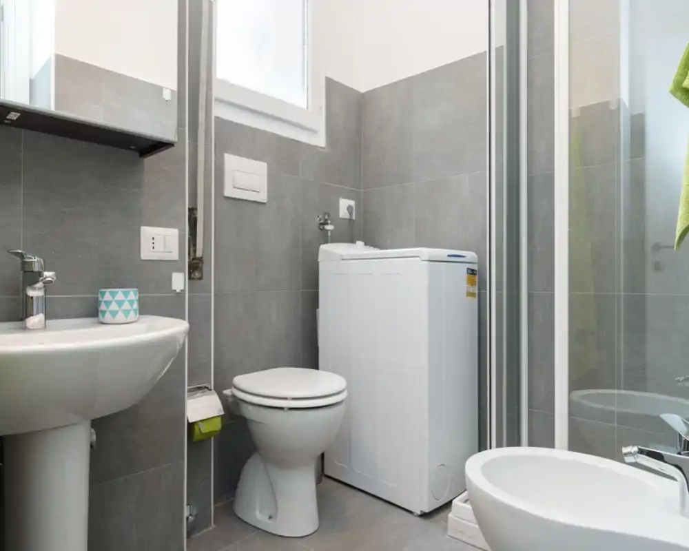 Compact modern bathroom with grey tiles, pedestal sink, toilet, bidet and a washing machine next to a glass shower enclosure. Bright, clean and functional layout suitable for a small apartment.