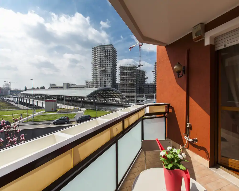 Sunny balcony with a small table and chair, modern frosted-glass railing and a view over nearby buildings and a transport hub — bright and inviting outdoor space.