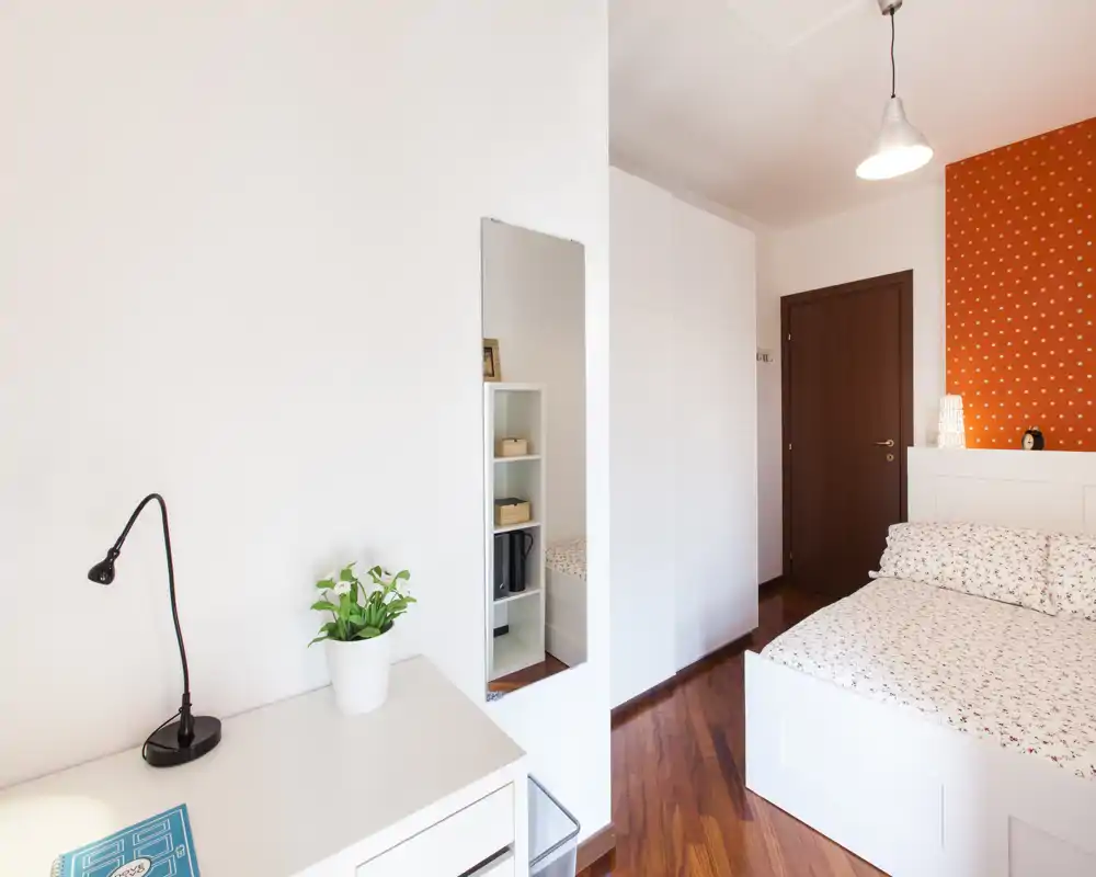 Bright, modern bedroom with a white bed, patterned bedding and warm wooden floors; neatly staged with a desk, lamp and decorative plant.