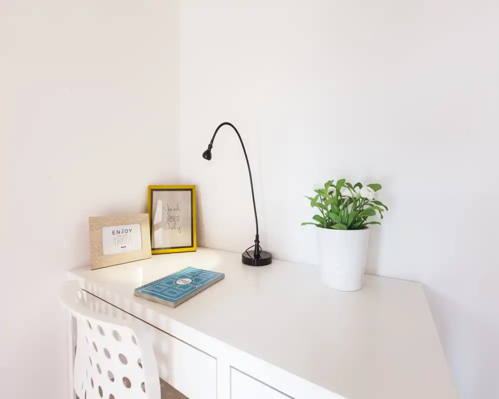Bright, minimal workspace corner with a white desk, chair, small lamp, framed prints and a potted plant — ideal for a compact home office or study nook.