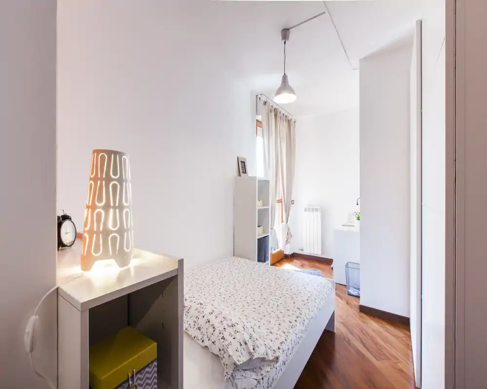 Bright, cozy bedroom with a double bed, patterned duvet, bedside table with lamp, and wooden floors—well lit by natural and artificial light.