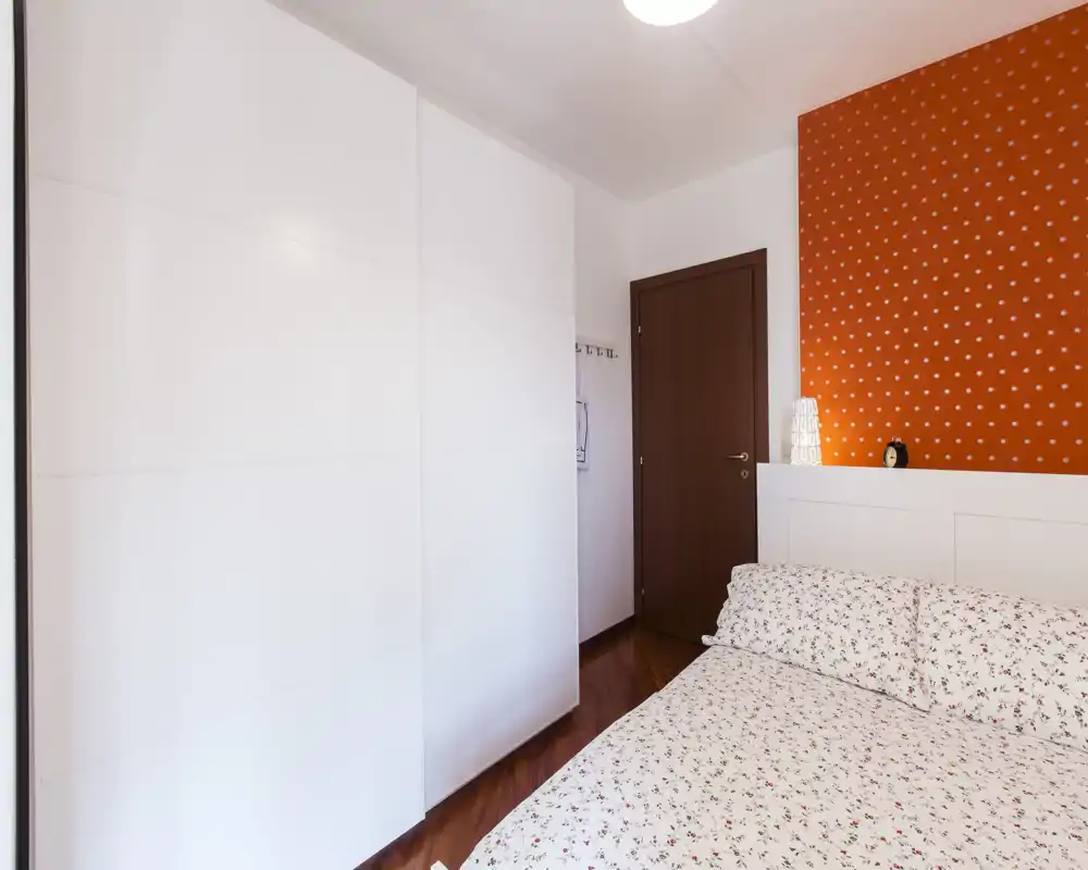 Bright and inviting bedroom with patterned bedding, a large white wardrobe and warm orange accent wall — ideal for a cozy stay.