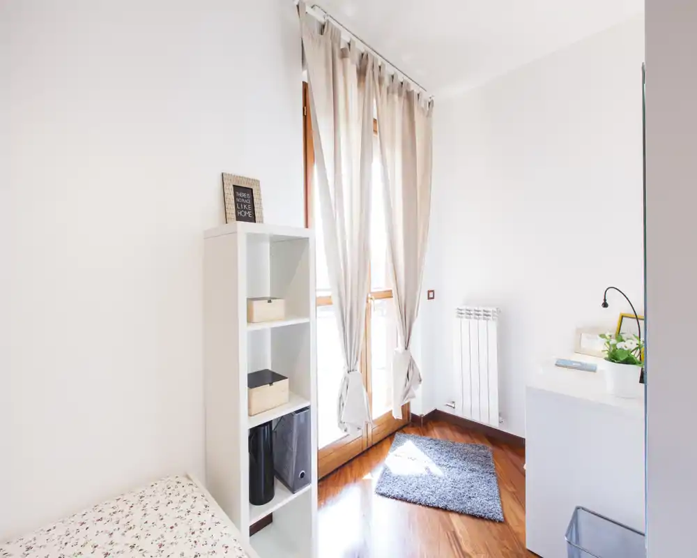 Bright, minimal bedroom corner with a single bed, shelving unit and large window letting in abundant natural light. The room features warm wooden floors, a small rug and simple, modern decor.