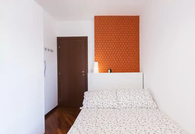 Dormitorio ordenado y luminoso con cama doble vestida con ropa de cama floral y suelo de madera cálida; un panel naranja con lunares y decoración sencilla crean un ambiente fresco y acogedor.
